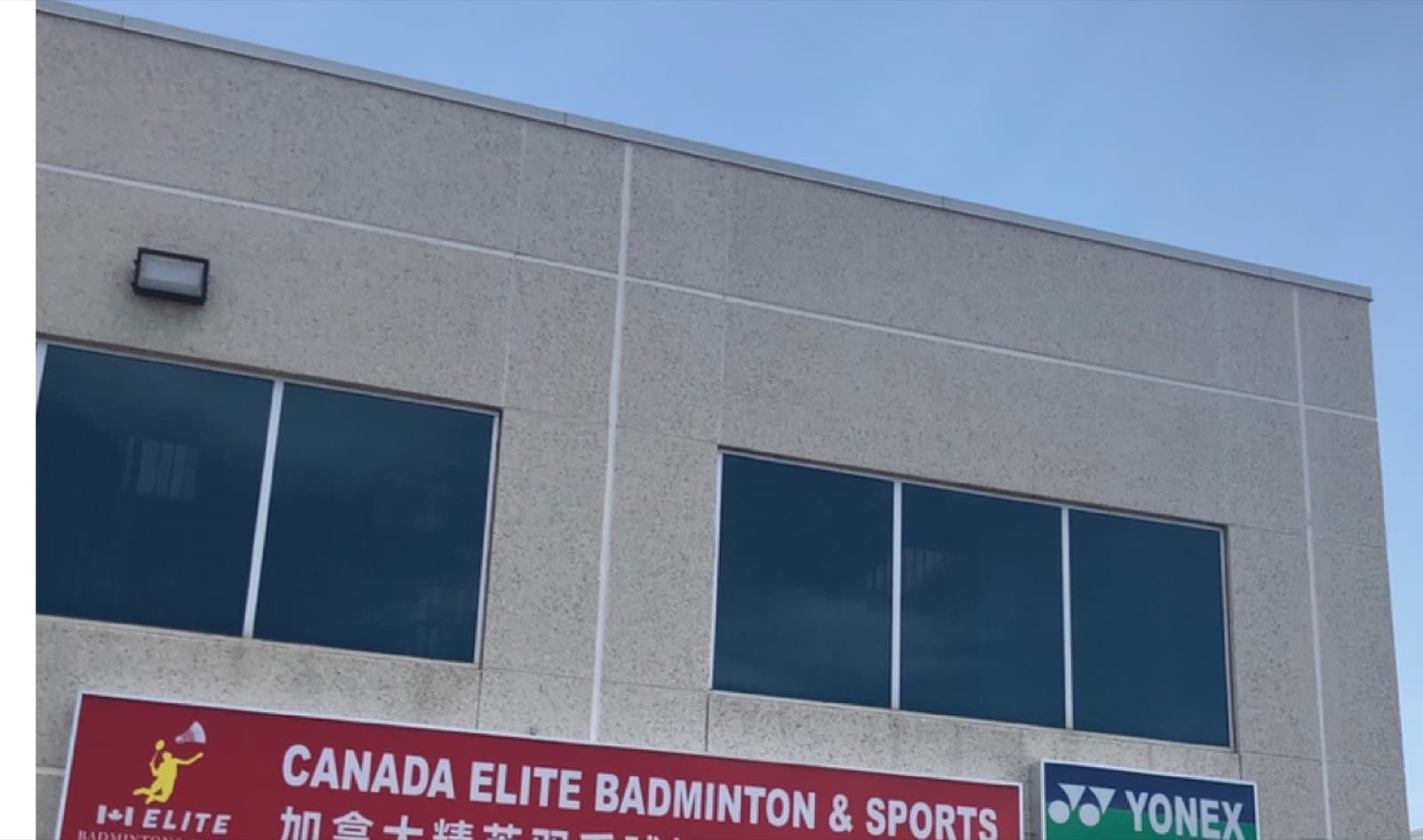 The exterior of Canada Elite Badminton's building, with the club's red sign and Yonex partner sign mounted on the wall under a clear sky.